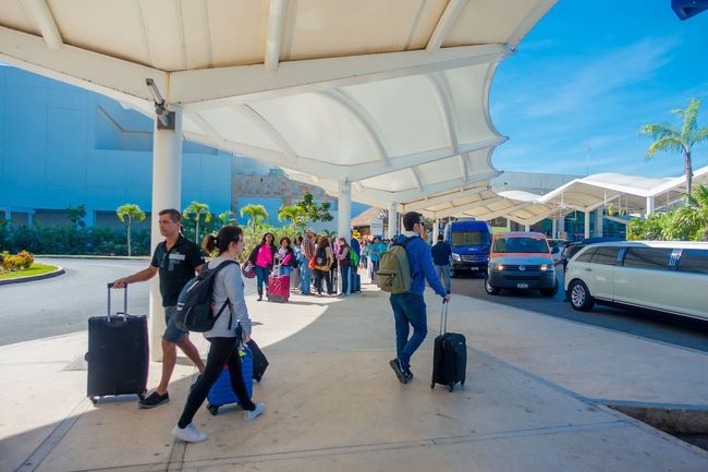 Viajero con maleta esperando en la banqueta del aeropuerto, revisando una app de transporte en el celular